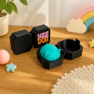 May include: A teal, rounded, ear-shaped fidget toy rests inside an open black hexagonal container. A second closed black hexagonal container features the text "NEE DOH" in pink and orange letters on its side.