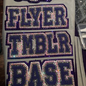 May include: A light gray leather sleeve featuring purple fabric patches with rhinestone borders. The patches display the text CAPTAIN, FLYER, TMBLR, and BASE in dark purple block letters, all outlined with shimmering iridescent rhinestones.