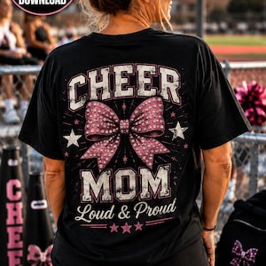 May include: A black short-sleeve t-shirt featuring a graphic on the back with the text CHEER MOM Loud & Proud. The design includes a large pink bow with a rhinestone pattern, white stars, and distressed lettering in white and pink colors.