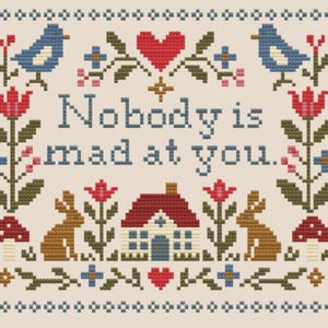 Puede incluir: Un patrón de punto de cruz sobre un fondo beige claro con el texto "Nobody is mad at you." El diseño incluye pájaros azules, corazones rojos, conejos marrones, casas con tejado rojo, setas rojas y motivos florales, enmarcado por un borde decorativo.