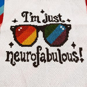 Op de afbeelding: Een wit stoffen vierkant met een kruissteekontwerp van een bril met zwarte randen en regenboogkleurige glazen. De tekst "I'm just neurofabulous!" is met zwart garen boven en onder de bril geborduurd, geaccentueerd door vier kleine zwarte sterretjes.