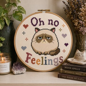 May include: A circular wooden embroidery hoop containing a cross-stitch design on off-white fabric. The design features a grumpy-faced cat in the center with the text "Oh no Feelings" above and below it, surrounded by small colorful hearts and stars.