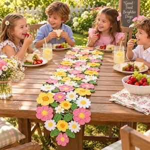 May include: A long, decorative table runner made of crocheted flowers in shades of white, yellow, and pink, accented with green crocheted leaves. The runner features a dense, interconnected floral pattern that spans the length of the wooden table.