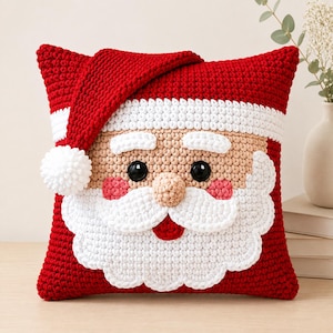 May include: A square crochet pillow designed to look like Santa Claus, featuring a red hat with a white pom-pom, a white brim, a beige face, black button eyes, pink cheeks, a beige nose, and a large white beard with a small red mouth.