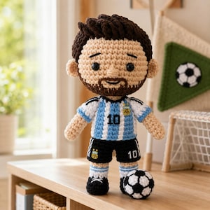 May include: A crochet doll of a soccer player with brown hair and a beard, wearing a light blue and white striped jersey with the number 10 and the Argentina national team crest, black shorts with the number 10, and a small crochet soccer ball.