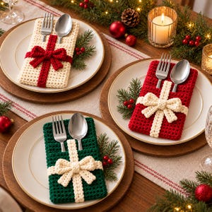 May include: Three cream, red, and green crocheted silverware holders shaped like gift boxes with bows. Each holder contains a silver fork and spoon, placed on white plates with gold rims, arranged on a wooden table with pine branches.