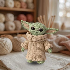 May include: A small, crocheted figure of a creature with large, dark, glossy eyes, pointed ears with pink inner detailing, and a light green head. The figure wears a textured, beige, long-sleeved robe with a prominent collar and vertical stitching.