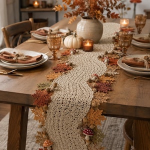 May include: A beige crocheted table runner featuring an intricate wavy pattern, decorated with crocheted autumn leaves in shades of orange, brown, and green, along with small crocheted mushrooms and acorns attached along the edges of the runner.