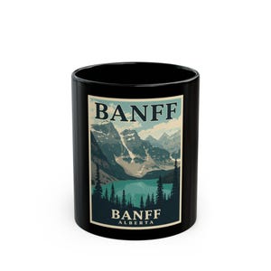 May include: A black ceramic mug featuring a rectangular graphic print of a mountain landscape with a lake and pine trees. The text BANFF is printed in large dark letters at the top, and BANFF ALBERTA is printed in smaller letters at the bottom.