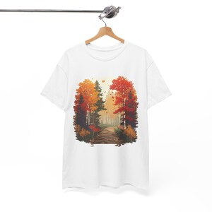 Könnte beinhalten: Ein weißes Kurzarm-T-Shirt mit einem zentralen Grafikdruck einer herbstlichen Waldszene. Die Illustration zeigt einen Feldweg, der sich durch Bäume mit leuchtend orangefarbenem, rotem und gelbem Laub unter einem hellen Himmel schlängelt.