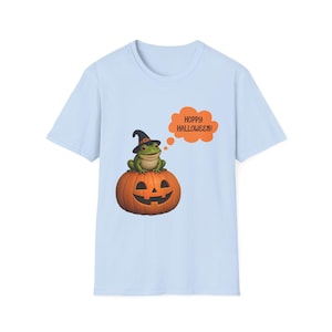 Puede incluir: Una camiseta azul claro con un gráfico de una rana verde con un sombrero de bruja negro, sentada sobre una calabaza naranja. Un globo de texto naranja junto a la rana contiene el texto en negro, HOPPY HALLOWEEN!