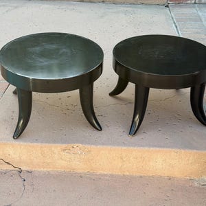 May include: Two dark brown, round side tables with curved legs. The tables have a circular top and a dark finish. The legs are curved and splayed, giving the tables a unique, modern look. The tables are placed on a concrete surface.