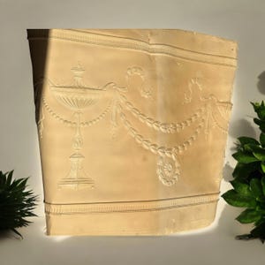 May include: A beige plaster mold of a decorative design featuring a vase with a floral motif, a garland of leaves, and a ribbon.