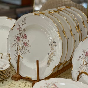 May include: A set of white porcelain dinner plates with a delicate floral design and gold trim. The plates are stacked on a wooden stand.