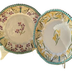May include: Two decorative ceramic plates with a scalloped edge and a blue and green rim. The plates feature a white background with a floral and butterfly design. One plate has a yellow floral design and the other has a purple floral design.