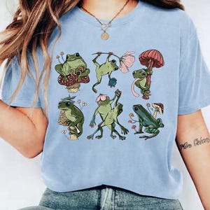 Può includere: Una t-shirt azzurra con una stampa grafica di sei rane verdi in varie pose. Le rane sono raffigurate mentre interagiscono con funghi, fiori e bacche, inclusa una rana seduta su un fungo e altre che eseguono movimenti di danza.