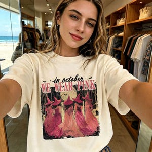 以下が含まれることがあります： クリーム色の半袖Tシャツ。森の中に立つピンク色の魔女の帽子とドレスを身にまとった5人の人物のグラフィックプリントが施されています。イラストの上部には黒とピンクの文字で「in october WE WEAR PINK」と書かれています。