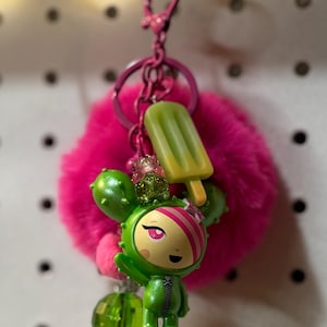 May include: A keychain featuring a bright pink faux fur pom-pom, a green cactus-themed character figurine with pink hair stripes, a light green popsicle charm, and a translucent green faceted bead, all attached to a pink metal lobster clasp ring.