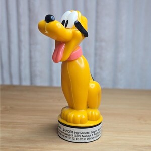 May include: A yellow plastic Pluto figurine with a black nose and ears, a pink tongue, and a pink collar. The character is mounted on a circular white base featuring printed text that lists ingredients and includes the Disney copyright mark.