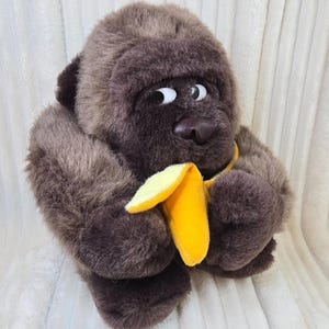 May include: A plush gorilla toy with dark brown fur, a rounded head, and large white eyes with black pupils looking to the side. The gorilla is holding a bright yellow plush banana in its hands, featuring a smooth texture and a curved shape.