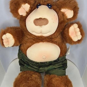 May include: A brown plush bear toy with a soft, light-coloured fabric face, blue eyes, and a tuft of brown hair on its head. The bear has a round belly with a visible navel and is wearing green camouflage-patterned shorts with an elastic waistband.