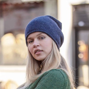 May include: A woman wearing a green jumper with a blue beanie and blue jeans. The jumper has white and red stripes on the cuffs.