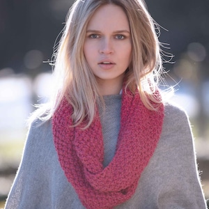 May include: A woman wearing a gray poncho with a pink knitted scarf. The poncho has white trim on the sleeves.