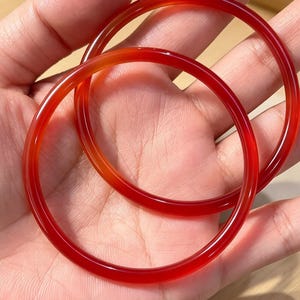 May include: Two smooth, circular bangles crafted from translucent red stone. The bracelets feature a polished, glossy finish that catches the light, showcasing a vibrant, deep crimson hue throughout the solid, continuous rounded bands.
