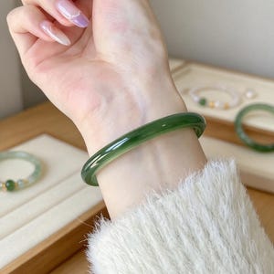 May include: A smooth, polished, dark forest green bangle bracelet with a translucent quality. The circular band features a rounded profile and a glossy finish, showcasing a deep, uniform green hue throughout the solid, continuous material.