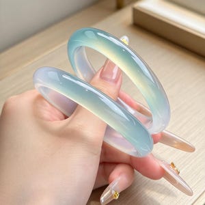 Puede incluir: Dos brazaletes rígidos lisos, translúcidos y de color azul verdoso claro con una superficie pulida y redondeada. Los brazaletes presentan una textura clara y vítrea que permite el paso de la luz, creando variaciones sutiles de color y profundidad.