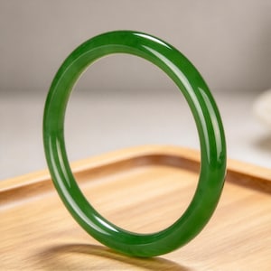May include: A smooth, circular bangle bracelet crafted from a solid piece of deep green stone. The bangle features a polished, glossy finish that reflects light, showcasing a consistent, vibrant emerald hue throughout its entire rounded form.