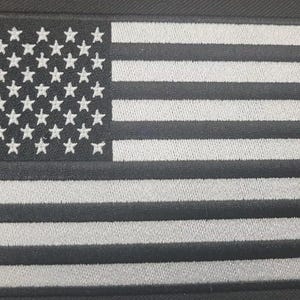 May include: A rectangular embroidered patch featuring the United States flag in a monochromatic black and grey color scheme. The design includes a field of stars on the left and horizontal stripes on the right, all rendered in stitched thread.