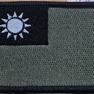 May include: A rectangular embroidered patch featuring a black canton in the upper left corner with a white twelve-rayed sun emblem. The remainder of the patch is an olive green color, and the entire border is finished with black stitching.