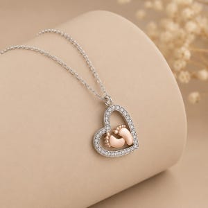 May include: A silver-toned necklace featuring a heart-shaped pendant. The heart frame is lined with small clear stones and contains a pair of small, smooth, rose-gold-toned baby feet positioned in the center of the open heart design.