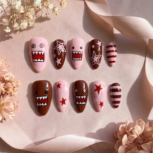 May include: A set of ten press-on nails featuring a mix of brown and light pink base colors. Designs include cartoon faces with jagged teeth, white and red star patterns, and horizontal stripes. The nails have a smooth, glossy finish throughout.