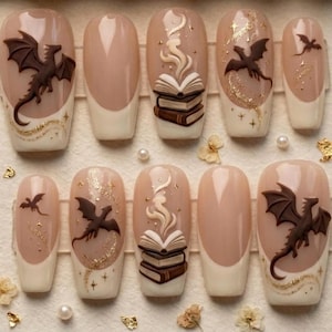 May include: A set of ten press-on nails featuring a nude base with cream-colored tips. The designs include raised brown dragon silhouettes, stacks of books with swirling smoke, and gold glitter accents in the shape of stars and flowing trails.