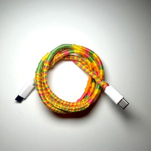 Può includere: Un cavo di ricarica USB-C avvolto a cerchio, caratterizzato da un rivestimento decorativo in filo giallo, rosa e verde brillante. Le estremità del cavo sono rifinite con connettori USB-C in plastica bianca standard su entrambi i lati.