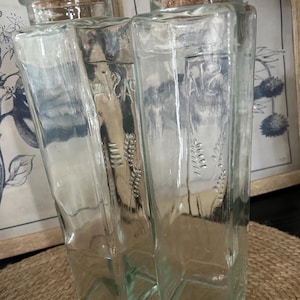 Peut inclure: Deux bouteilles rectangulaires hautes en verre transparent avec des bouchons en liège. Chaque bouteille présente le texte en relief PASTA et un motif décoratif d'épi de blé sur la face avant. Les récipients ont une base carrée.