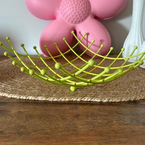 Peut inclure: Une coupe à fruits en fil métallique vert citron dotée d'une structure en grille avec des rayons incurvés vers le haut, chacun surmonté d'une petite pointe sphérique. La coupe est faite de fines tiges métalliques disposées en croisillons.