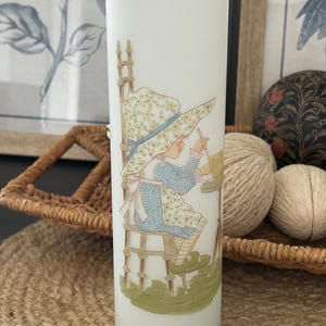 May include: A tall, cylindrical frosted glass vase featuring a gold-rimmed top edge. The exterior displays a printed illustration of a person wearing a wide-brimmed floral hat and a blue patterned apron, sitting on a chair while writing at a desk.