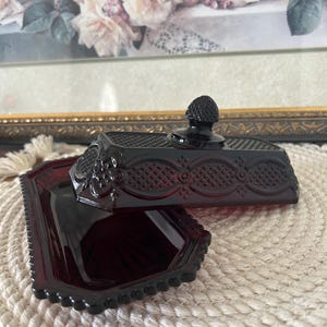 Peut inclure: Un beurrier en verre rouge profond composé d'une base rectangulaire et d'un couvercle assorti orné d'un motif en losanges texturé et d'un bouton central. Le verre présente une finition lisse et polie avec des détails en relief complexes.