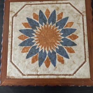 May include: A square ceramic tile trivet featuring a central floral pattern with alternating blue and orange petals. The tile is set within a dark brown wooden frame with a scalloped edge, showcasing a weathered, off-white background texture.