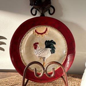 May include: A round ceramic decorative plate featuring a raised rooster figure in the centre, a sun motif above, and a deep red rim. The plate is held by a dark brown metal stand that includes a decorative rooster topper at the very top.