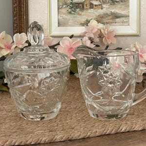 Peut inclure: Un sucrier en verre transparent avec couvercle assorti et un pot à lait, tous deux ornés d'un motif en étoile pressé et de détails géométriques à facettes. Le sucrier possède un bouton pointu sur le couvercle, tandis que le pot a une anse.