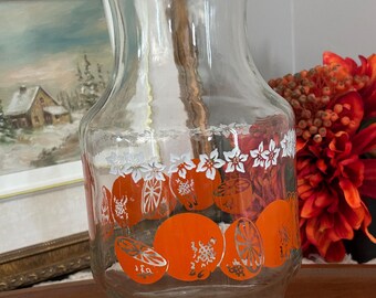 May include: A clear glass pitcher featuring a decorative band of white flowers and vibrant orange citrus fruit illustrations. The vessel has a wide base, a tapered neck, and a flared rim, showcasing a repeating pattern of whole and sliced oranges.