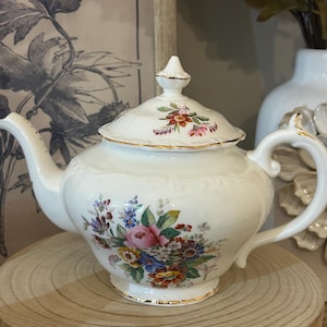 Peut inclure: Une théière en porcelaine de couleur crème ornée d'un bouquet floral délicat composé de roses roses, de myosotis bleus et de fleurs jaunes. La théière comprend un couvercle assorti avec un bouton pointu, des bords dorés et des détails en relief.