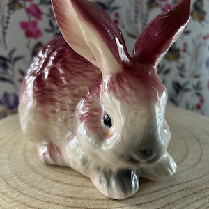 May include: A ceramic rabbit figurine featuring a glossy finish with a colour palette of deep reddish-pink and creamy white. The rabbit is depicted in a seated, alert posture with long, upright ears, dark eyes, and detailed fur texturing.