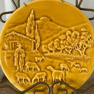 May include: A circular mustard yellow ceramic decorative plate featuring a raised relief scene of a shepherd standing near a stone cottage, tall cypress trees, and a flock of sheep grazing in a rolling landscape under a clear sky.