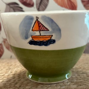 May include: A ceramic bowl with a white upper section and a green lower section. The white portion features a hand-painted illustration of an orange sailboat with a red sail, set against a light blue wash background and dark blue wavy water.