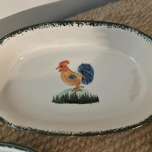 May include: An oval-shaped white ceramic serving dish featuring a hand-painted rooster illustration in the centre. The rooster has a red comb, tan body, and blue tail feathers. The rim has a dark green speckled finish along the edge.
